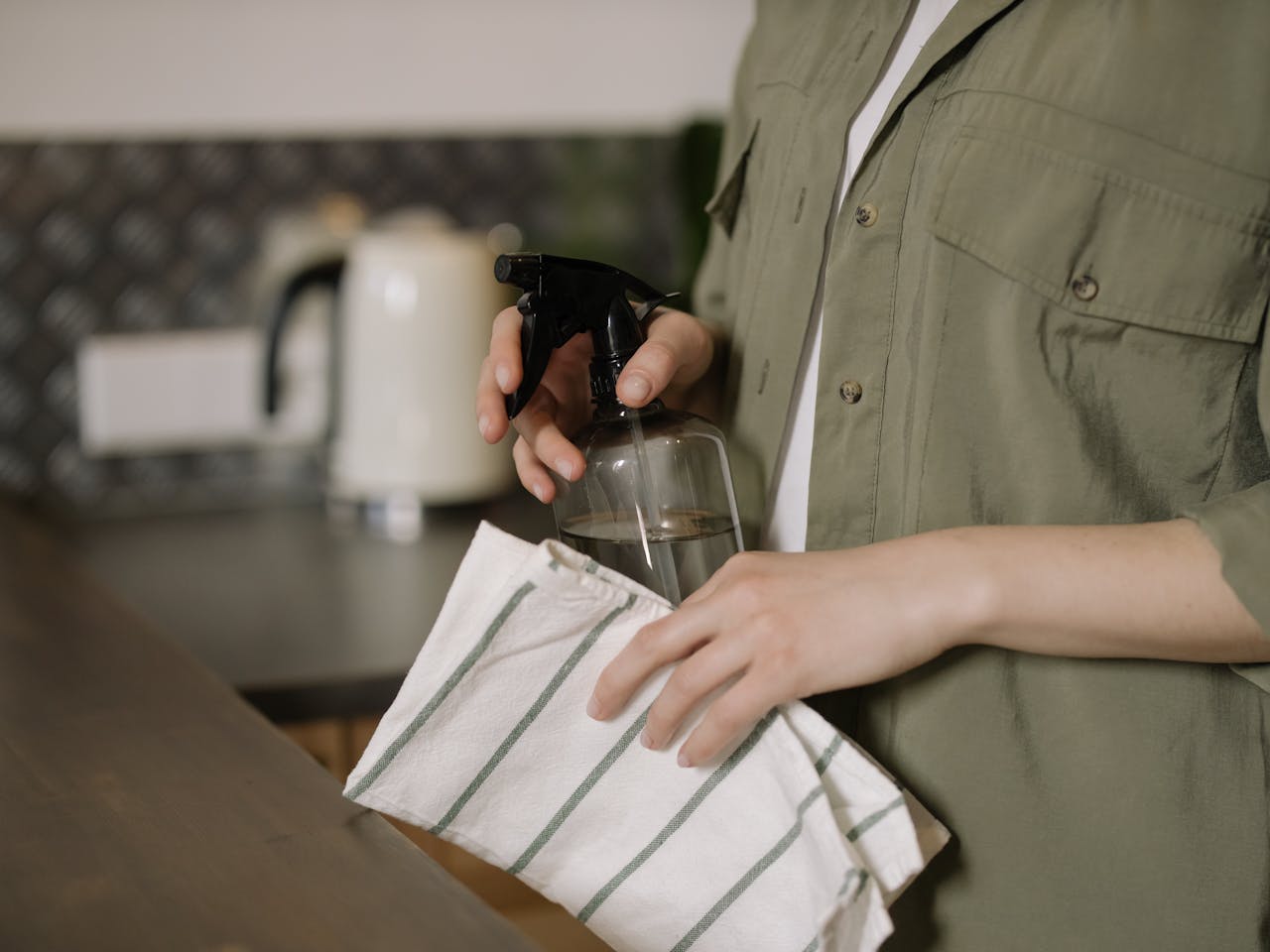 about-us-02 A person cleaning a kitchen counter with a spray bottle and cloth, maintaining cleanliness.